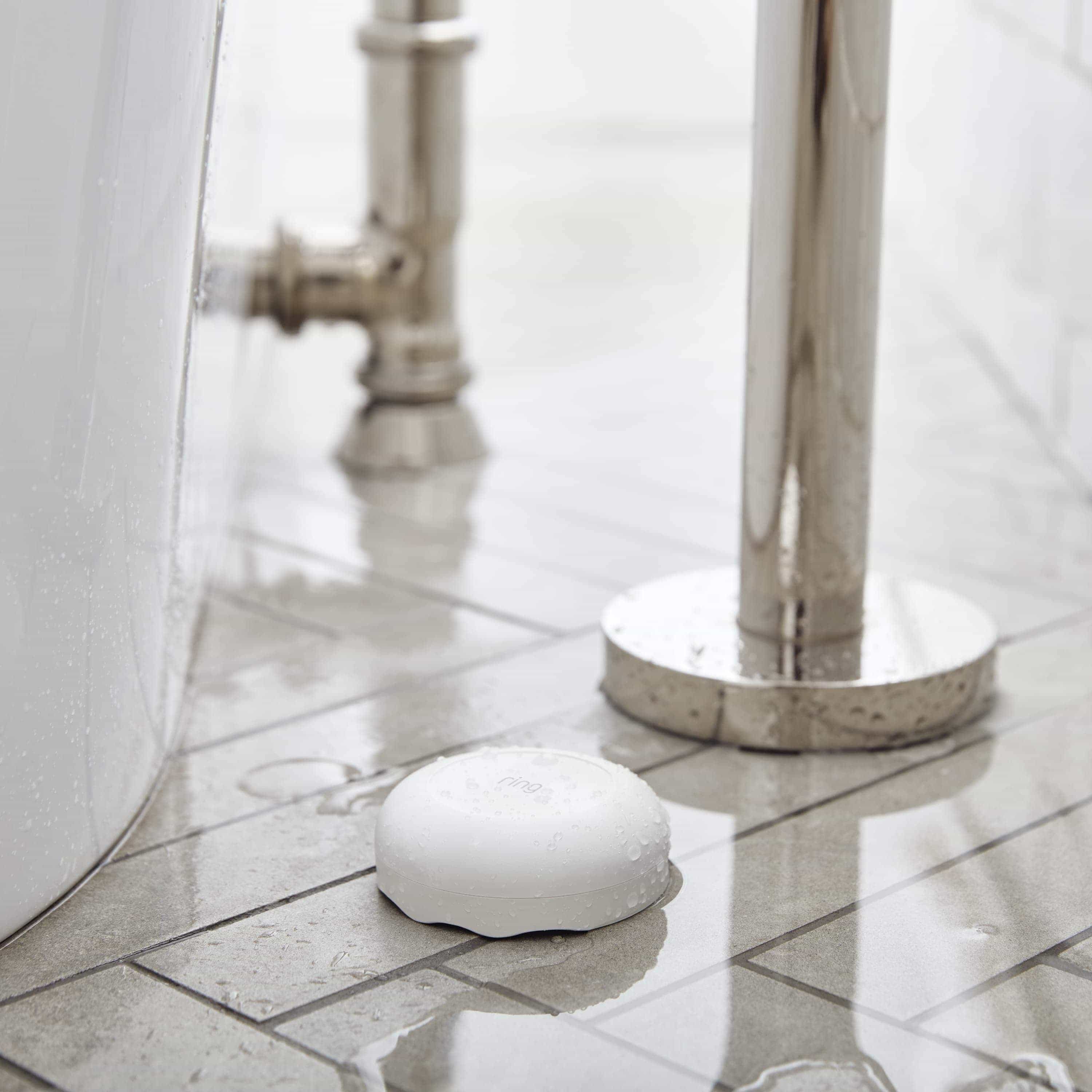 Flood & Freeze Alarm Bundle - White Ring Alarm Flood and Freeze Sensor placed on a wet, tiled floor next to a bathtub and plumbing fixtures.