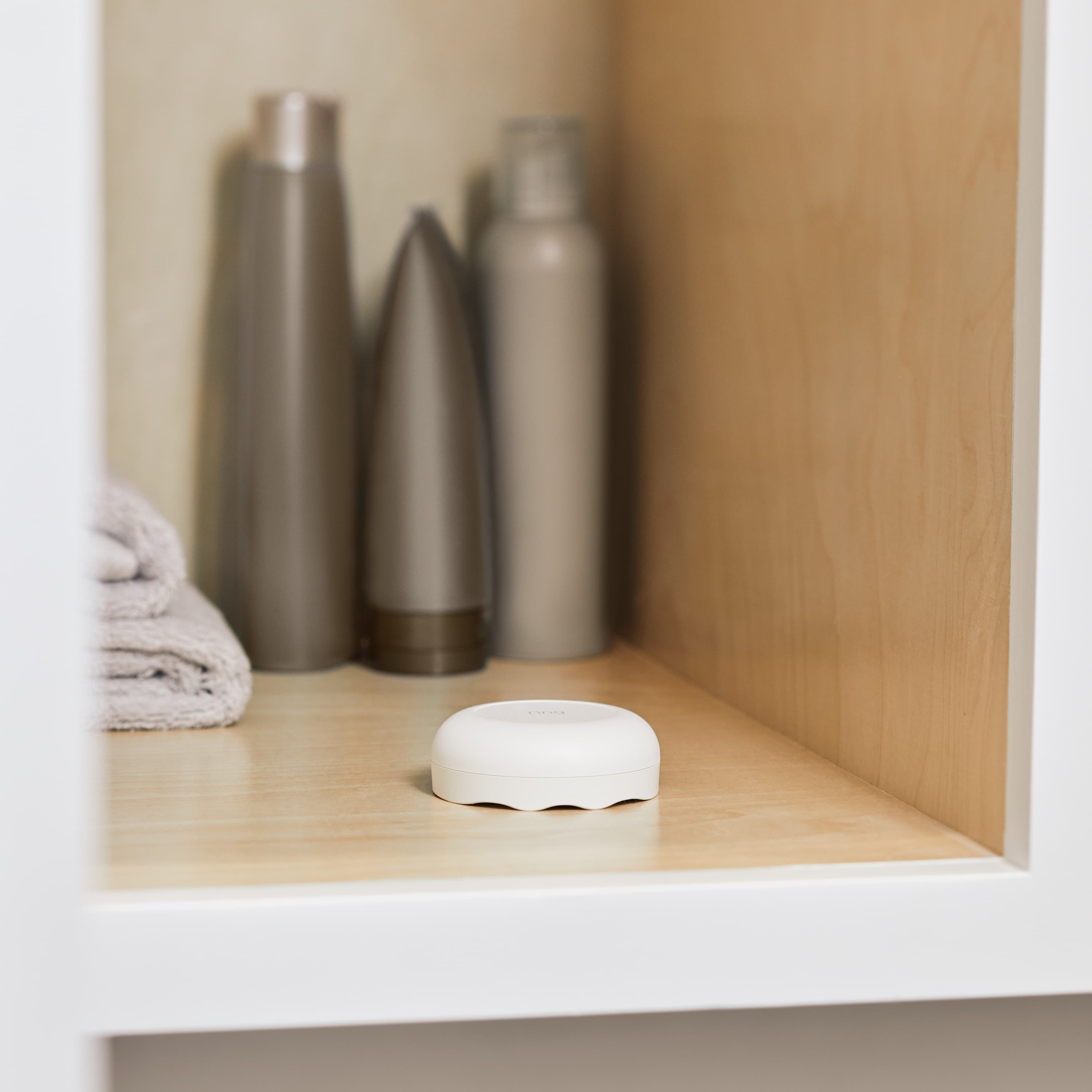 Flood & Freeze and Smoke & CO Listener Alarm Bundle - White Ring Alarm Flood and Freeze Sensor placed on a wooden shelf next to folded towels and toiletry bottles. 