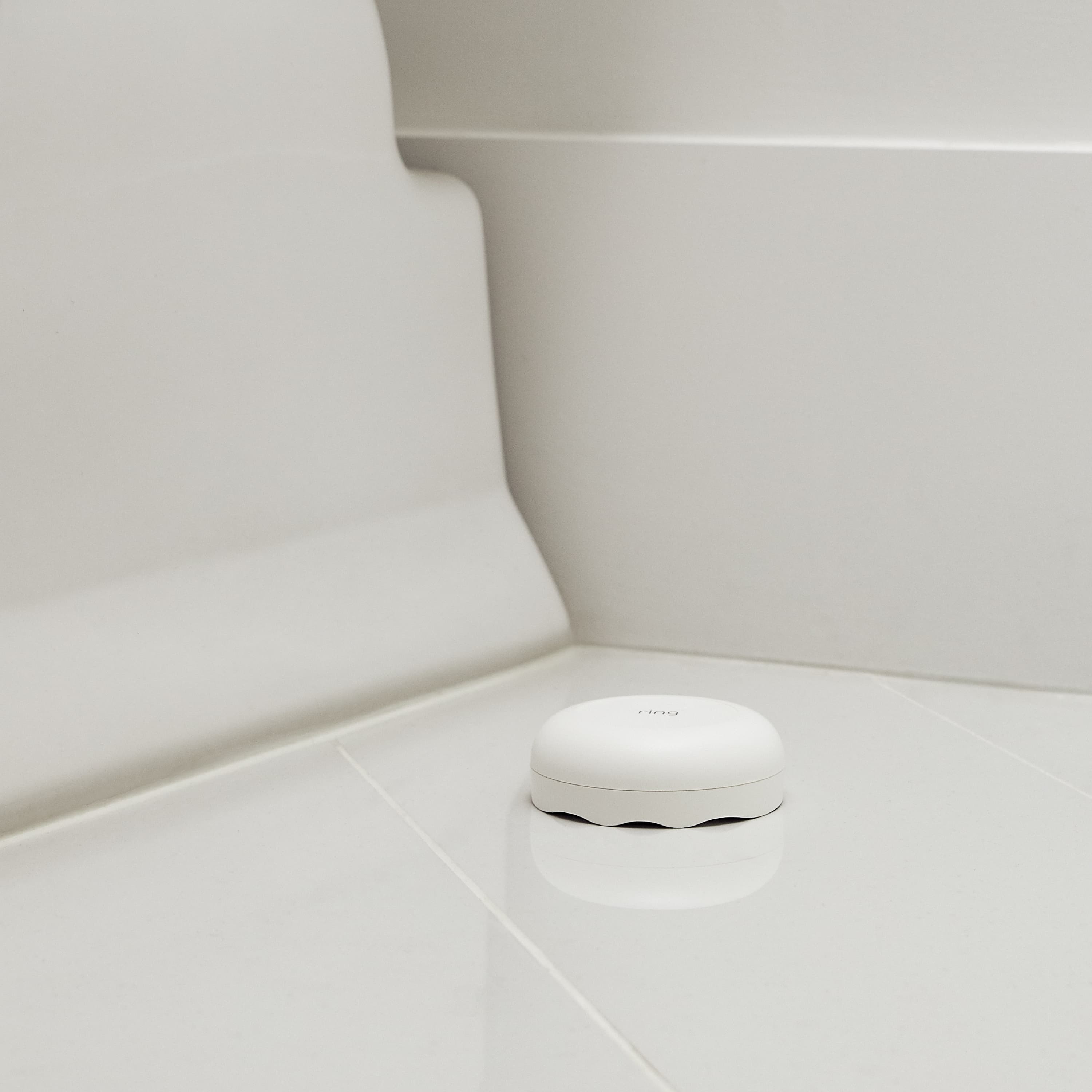 Flood & Freeze and Smoke & CO Listener Alarm Bundle - White Ring Alarm Flood and Freeze Sensor placed on the tiled bathroom floor near a bathtub. 