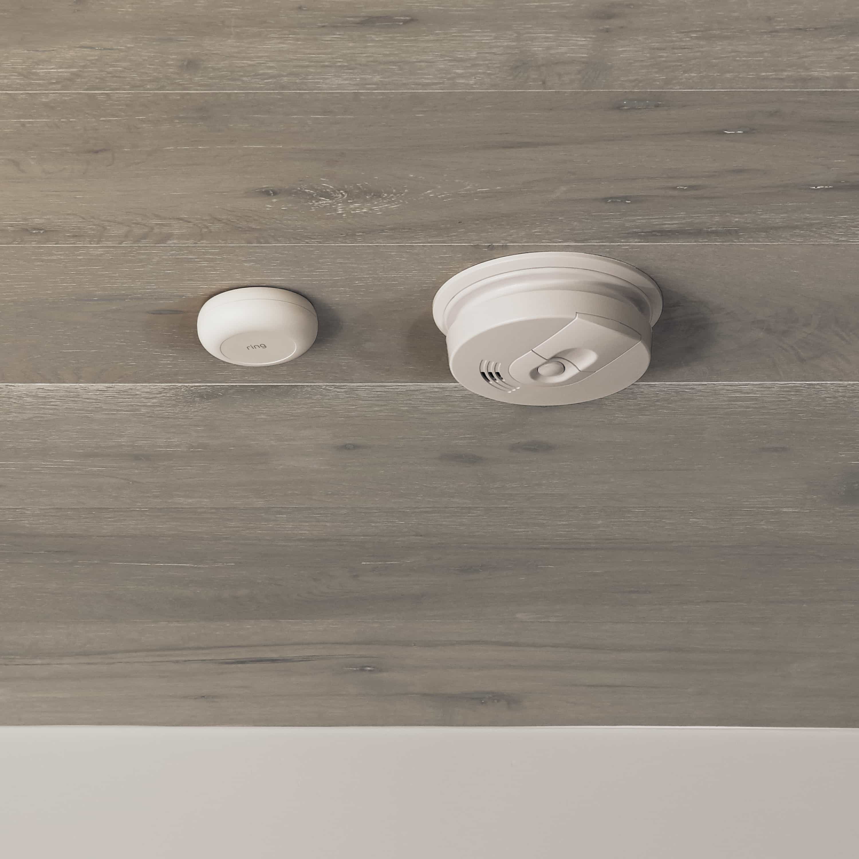 Flood & Freeze and Smoke & CO Listener Alarm Bundle - White Ring Alarm Smoke and CO Listener mounted on a wood-paneled ceiling next to a traditional smoke detector.
