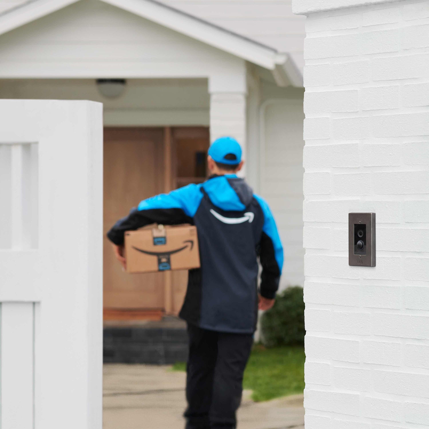 Video Doorbell Elite (Power over Ethernet) - Amazon delivery driver carrying package walks toward a house, near black Ring Video Doorbell Elite on white brick wall.
