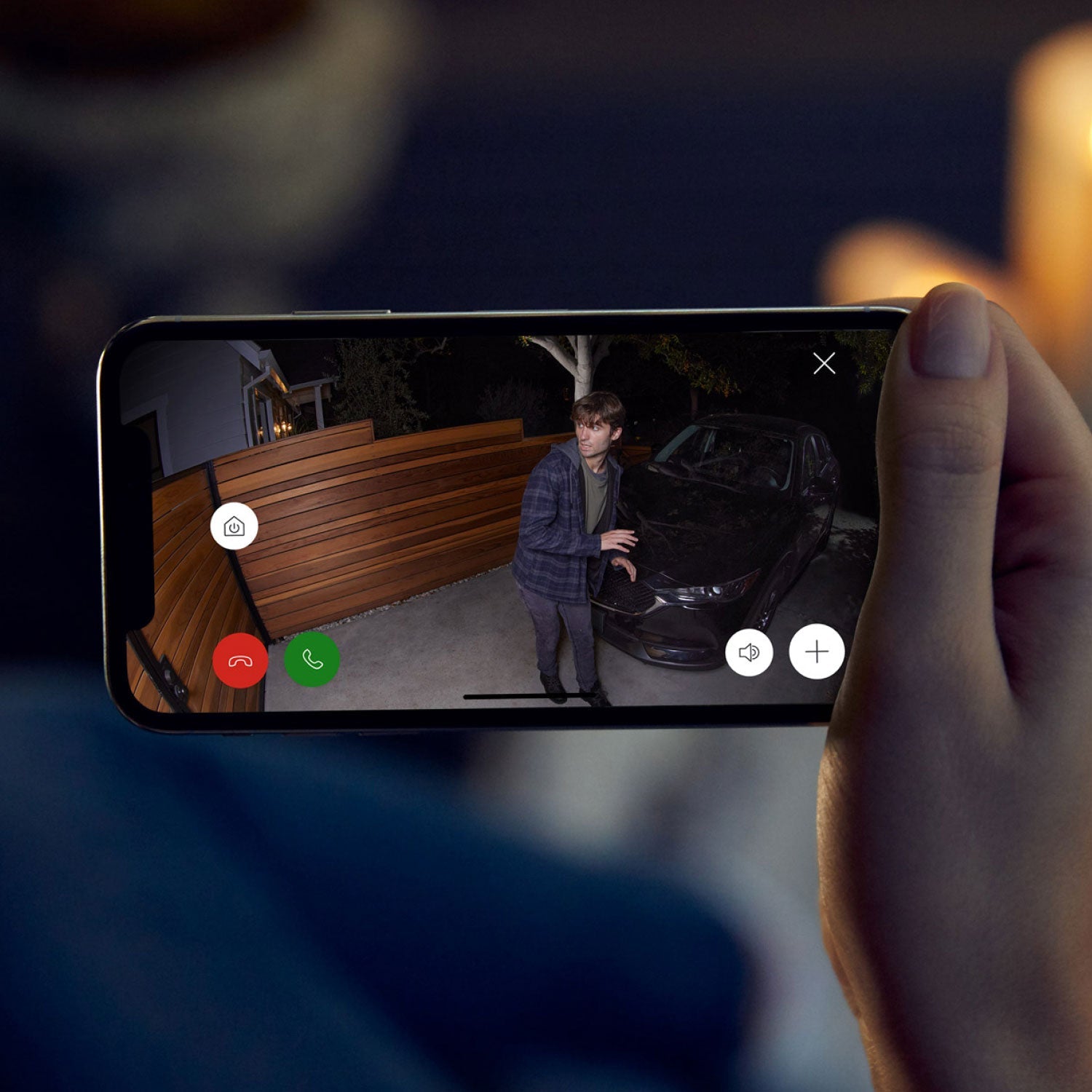 Spotlight Cam Plus - Wired:Hand holding a smartphone showing live Ring Spotlight Cam Plus video footage of a suspicious person near a car at night, with app controls visible on the screen.