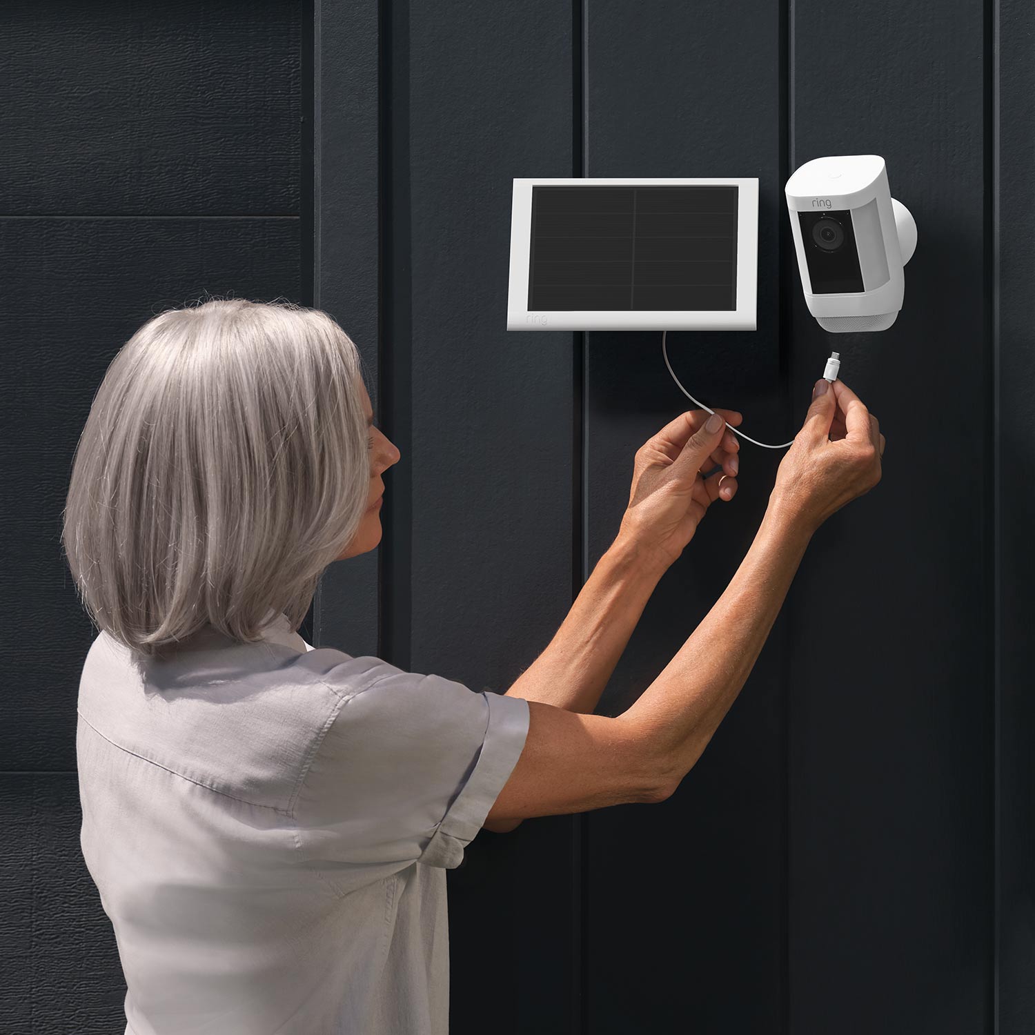 Solar Panel (USB-C) (for Outdoor Cam (Stick Up Cam), Outdoor Cam Plus, Stick Up Cam Pro, Spotlight Cam Plus, Spotlight Cam Pro) - Woman connecting a white Ring Solar Panel USB-C to a white Ring security camera mounted on a dark exterior wall.