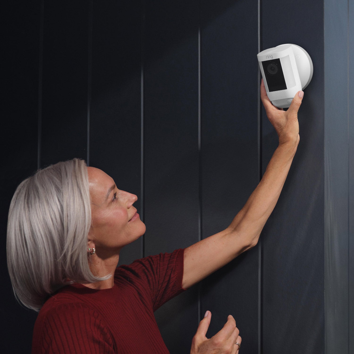 Spotlight Cam Pro (Featuring Retinal 2K) - Wired:Woman reaching up to adjust a white Spotlight Cam Pro mounted on a dark exterior wall of home.