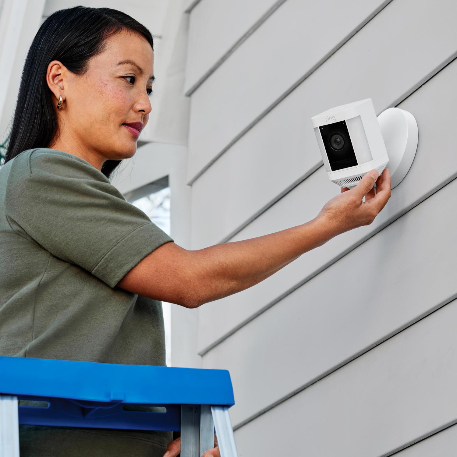 Spotlight Cam Plus - Wired:Woman on ladder adjusting a white Ring Spotlight Cam Plus mounted on a grey exterior wall with horizontal siding.