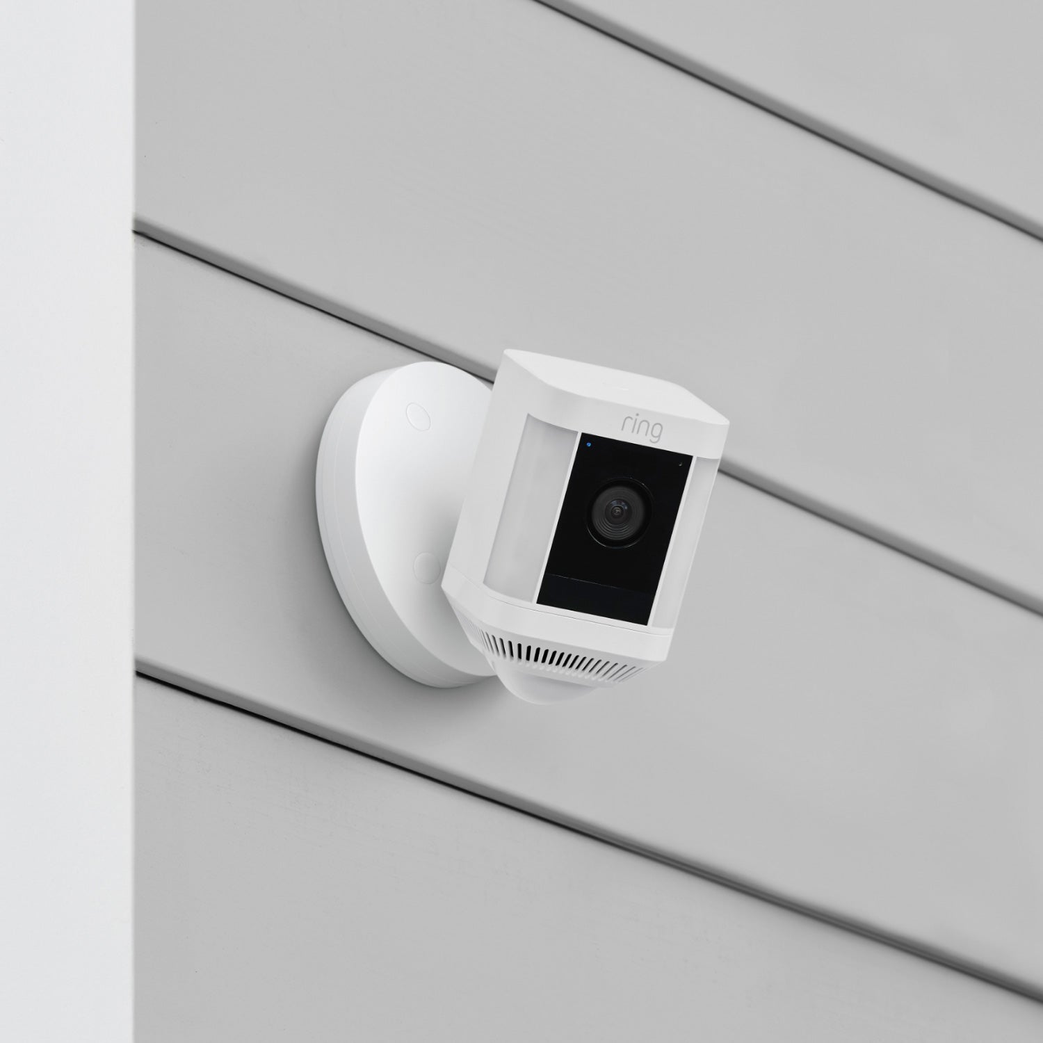 Spotlight Cam Plus - Wired:White Ring Spotlight Cam Plus mounted on a grey exterior wall with horizontal siding, shown angled downward.