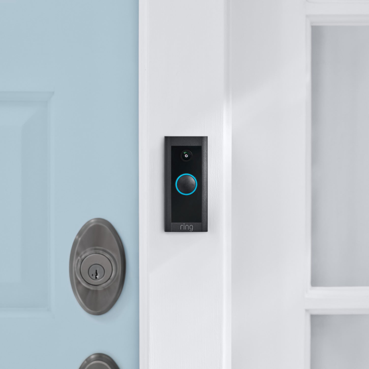 Video Doorbell Wired - Black Ring Video Doorbell Wired with blue-lit button mounted on white door frame next to a blue front door.