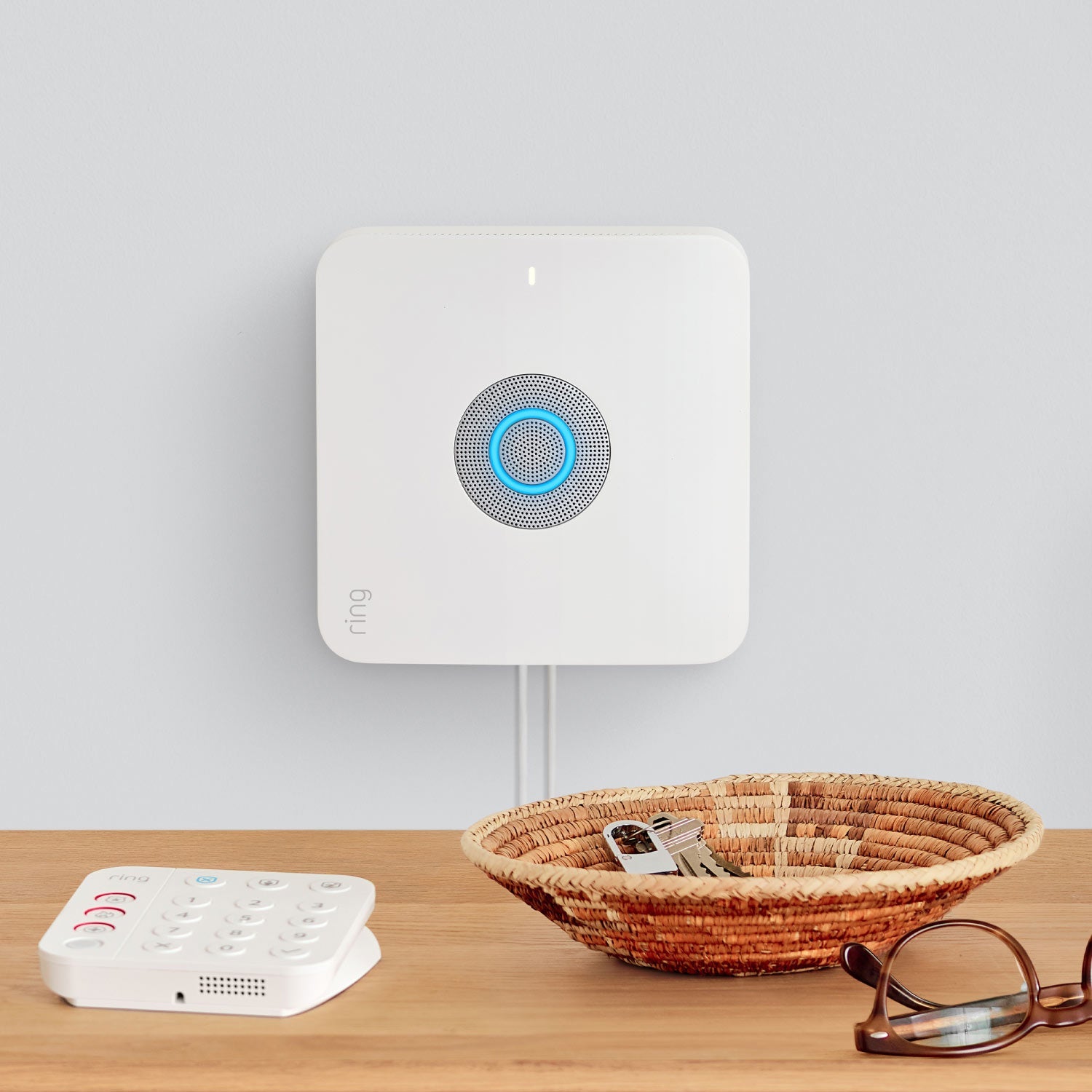 Protected Enhanced Kit (Battery Doorbell Plus + Alarm Security Kit, 8-Piece) - Wall-mounted Ring Alarm Base Station with glowing blue light next to tabletop Ring keypad, basket of keys, and glasses.