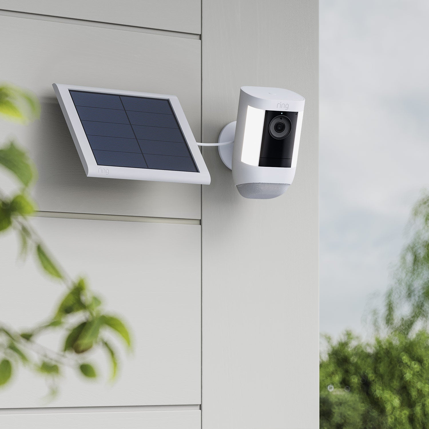 Spotlight Cam Pro (Featuring Retinal 2K) - Solar:White Ring Spotlight Cam Pro with lights on, mounted on an exterior wall and connected to a white Ring Solar Panel.