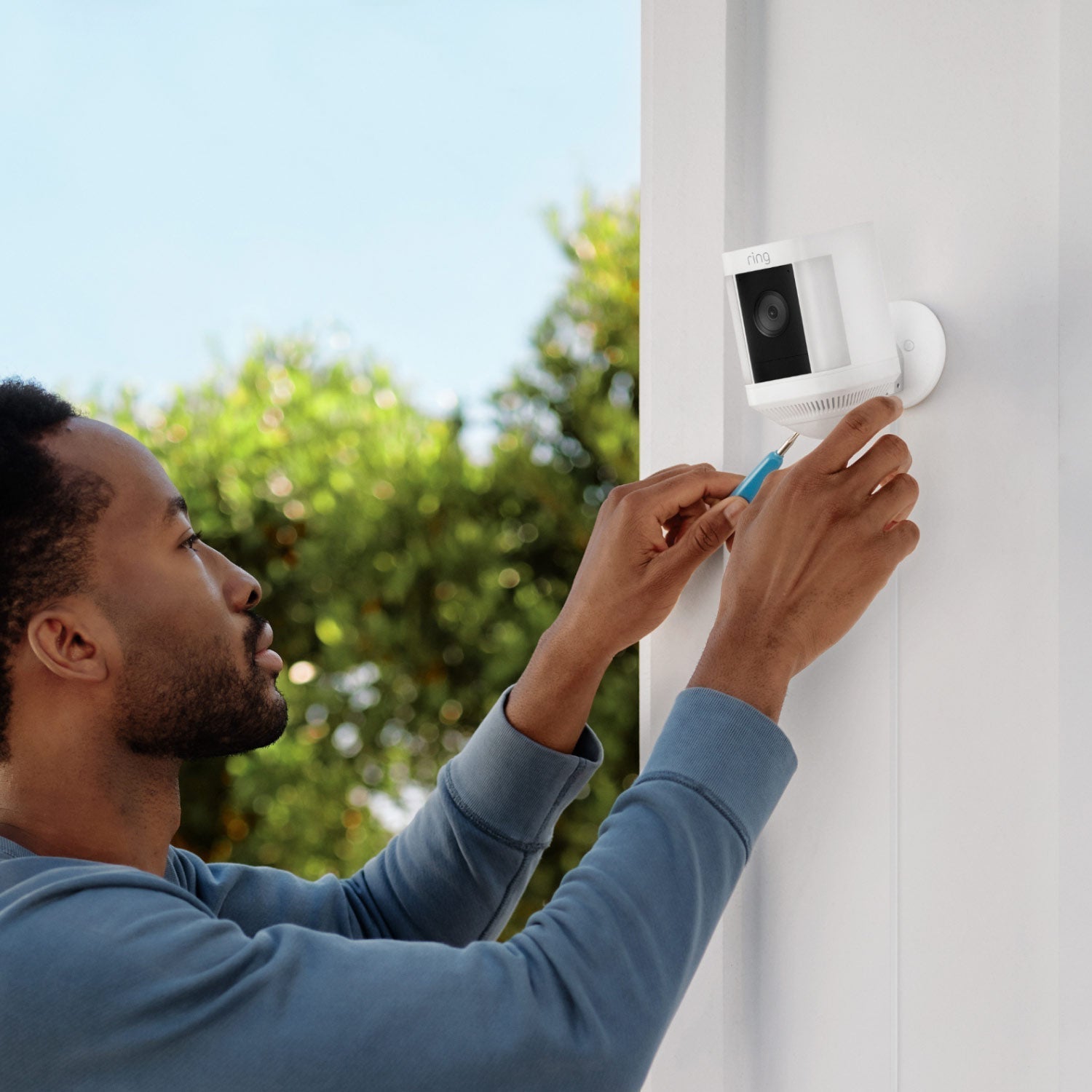 Spotlight Cam Plus - Plug-In:Man using a screwdriver to install a white Spotlight Cam Plus on an exterior white wall.