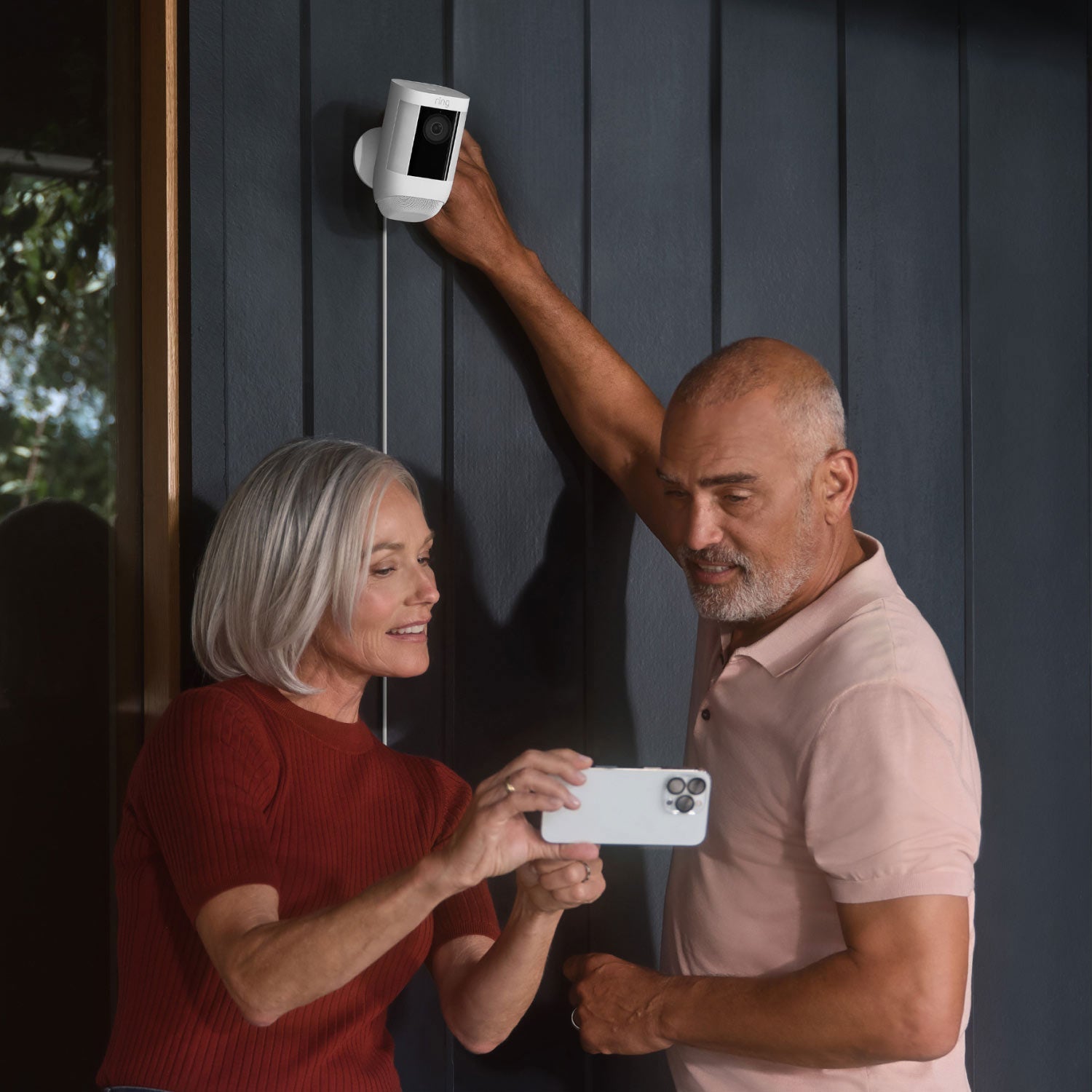 Spotlight Cam Pro (Featuring Retinal 2K) - Plug-In:Two people installing white Ring Spotlight Cam Pro on exterior wall of home, while checking Ring App settings.