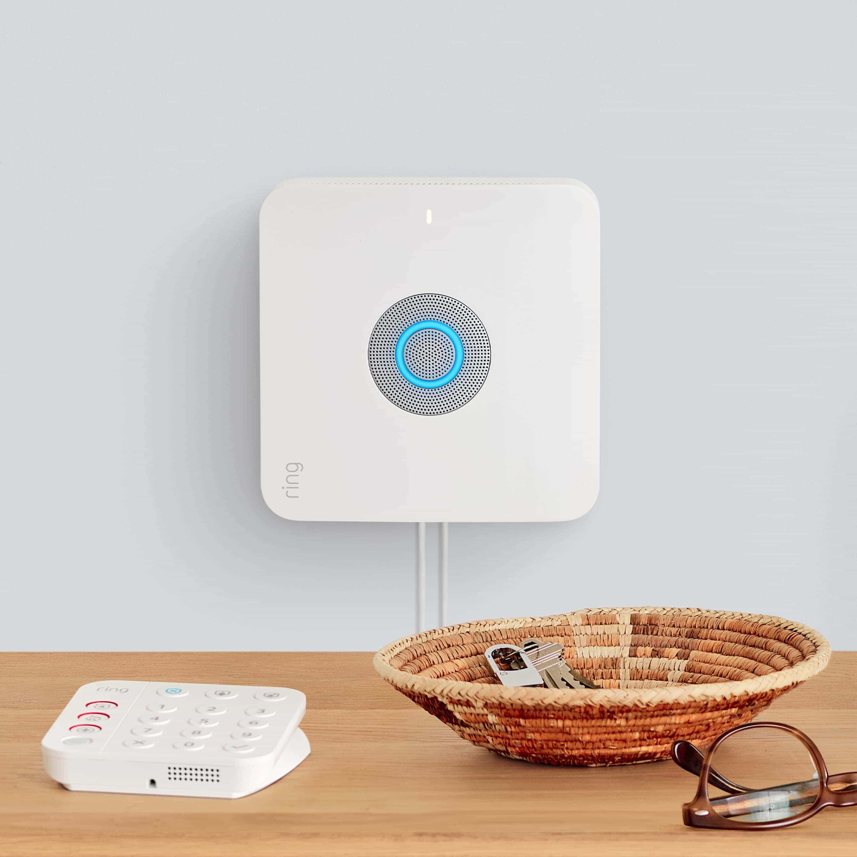 Alarm Security Kit, 8-Piece (Certified Refurbished) - wall-mounted Ring Alarm Base Station with glowing blue light, by tabletop Ring Alarm Keypad, basket of keys, and glasses.
