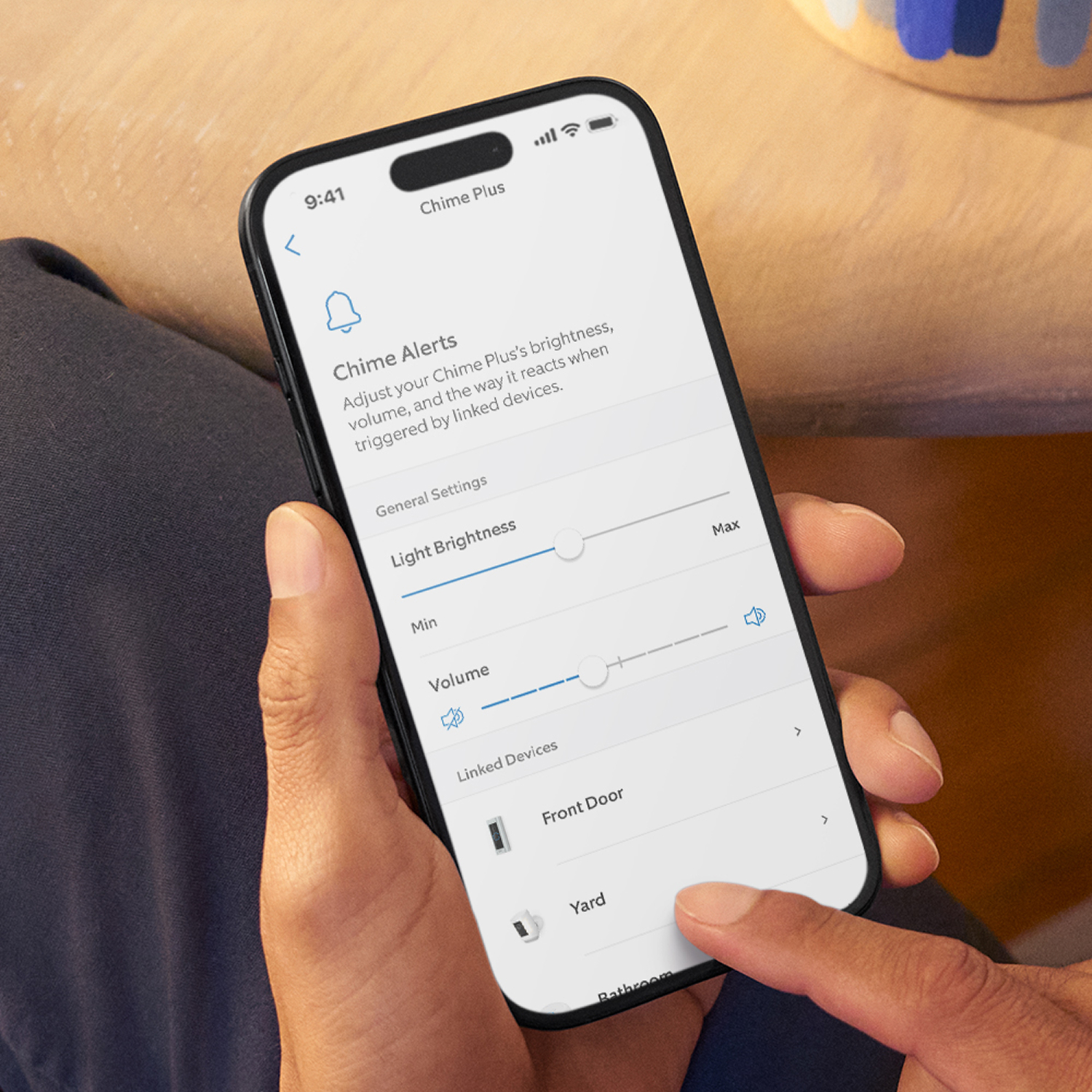 Chime Plus - Hand holding a smartphone showing a Chime Plus app screen with sliders for light brightness and volume, and linked devices list.