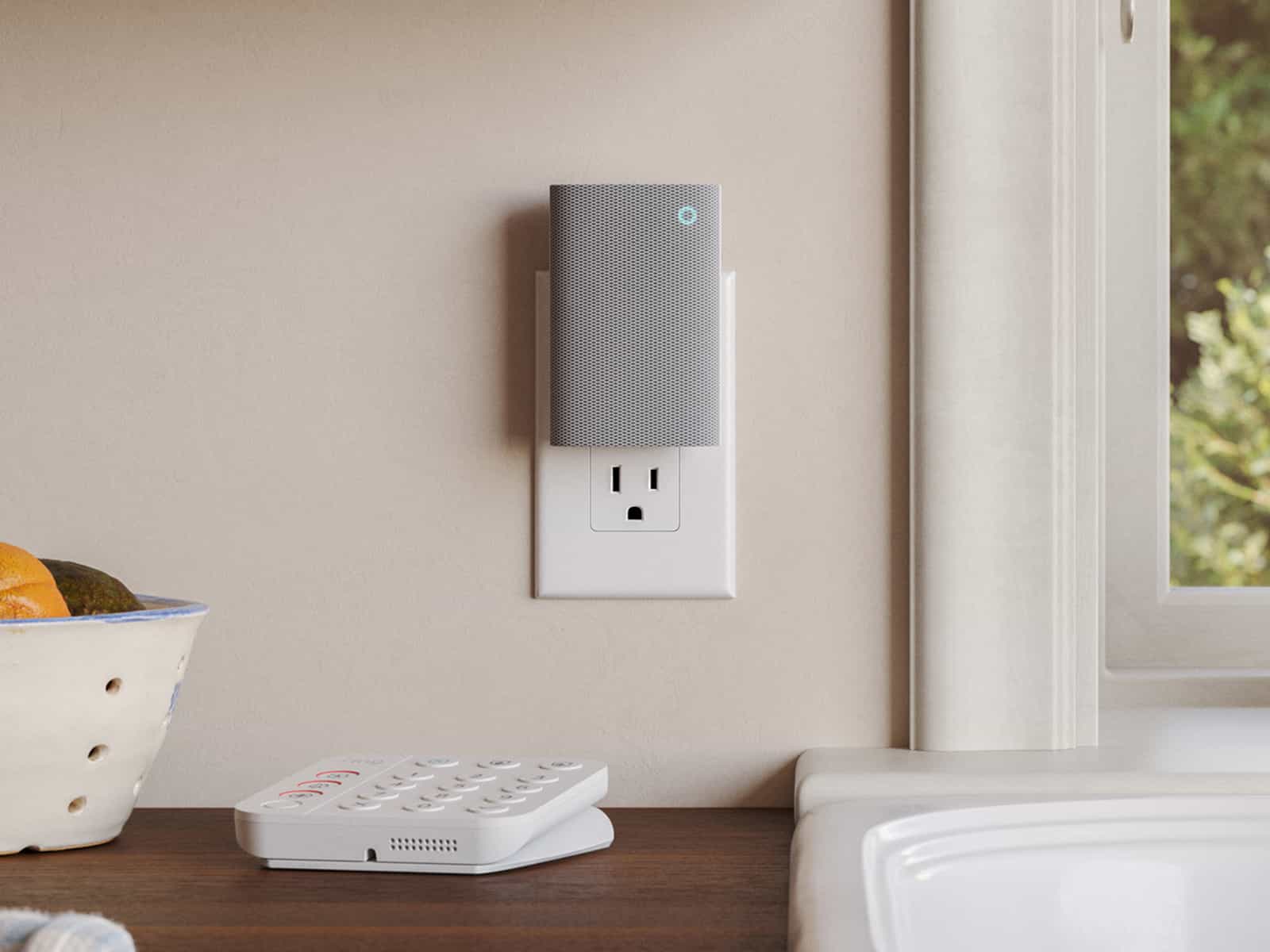 Gray Ring Chime Plus with blue light in wall outlet above kitchen counter, near window; white keypad sits on the counter.