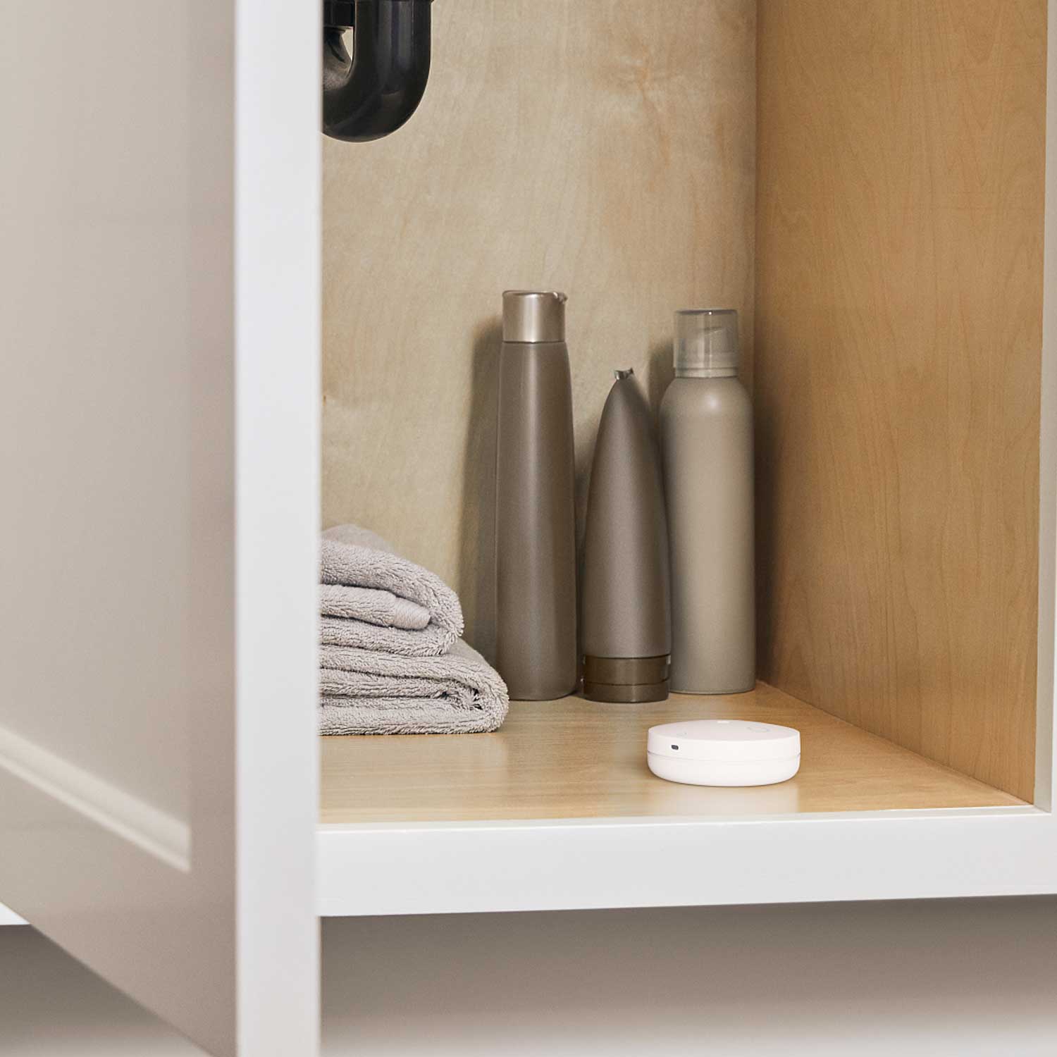 Flood & Freeze Sensor (Newest Model) - Bathroom shelf with folded gray towels, three metallic toiletry bottles, and a small white circular device.