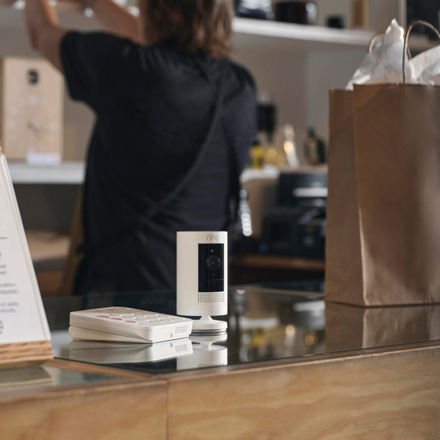 Outdoor Cam (Formerly: Stick Up Cam) - Battery:White Ring Outdoor Stick Up Cam next to keypad on a store counter, with person standing by shelves visible.