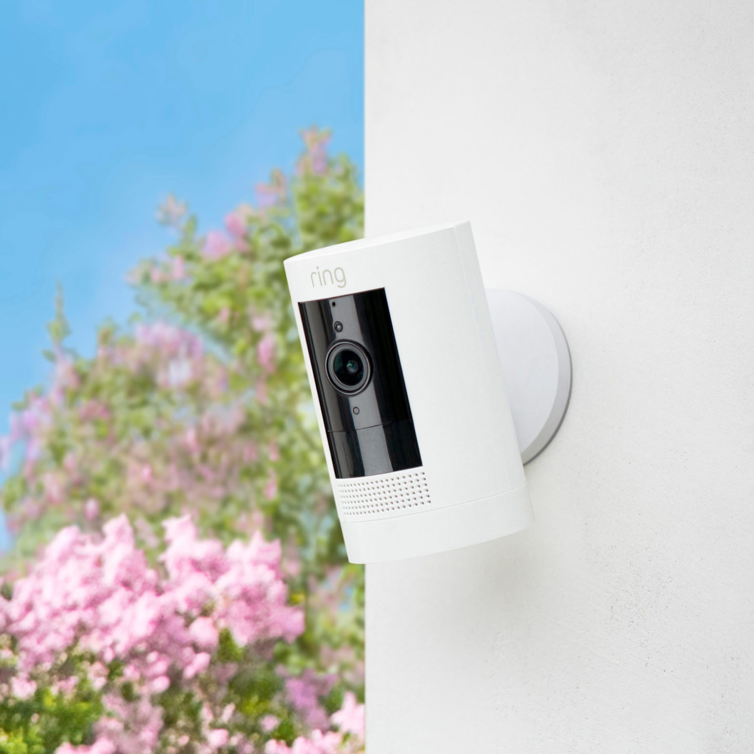 Outdoor Cam (Formerly: Stick Up Cam) - Battery:White Outdoor Cam mounted on a wall outdoors with trees and flowers in the background.