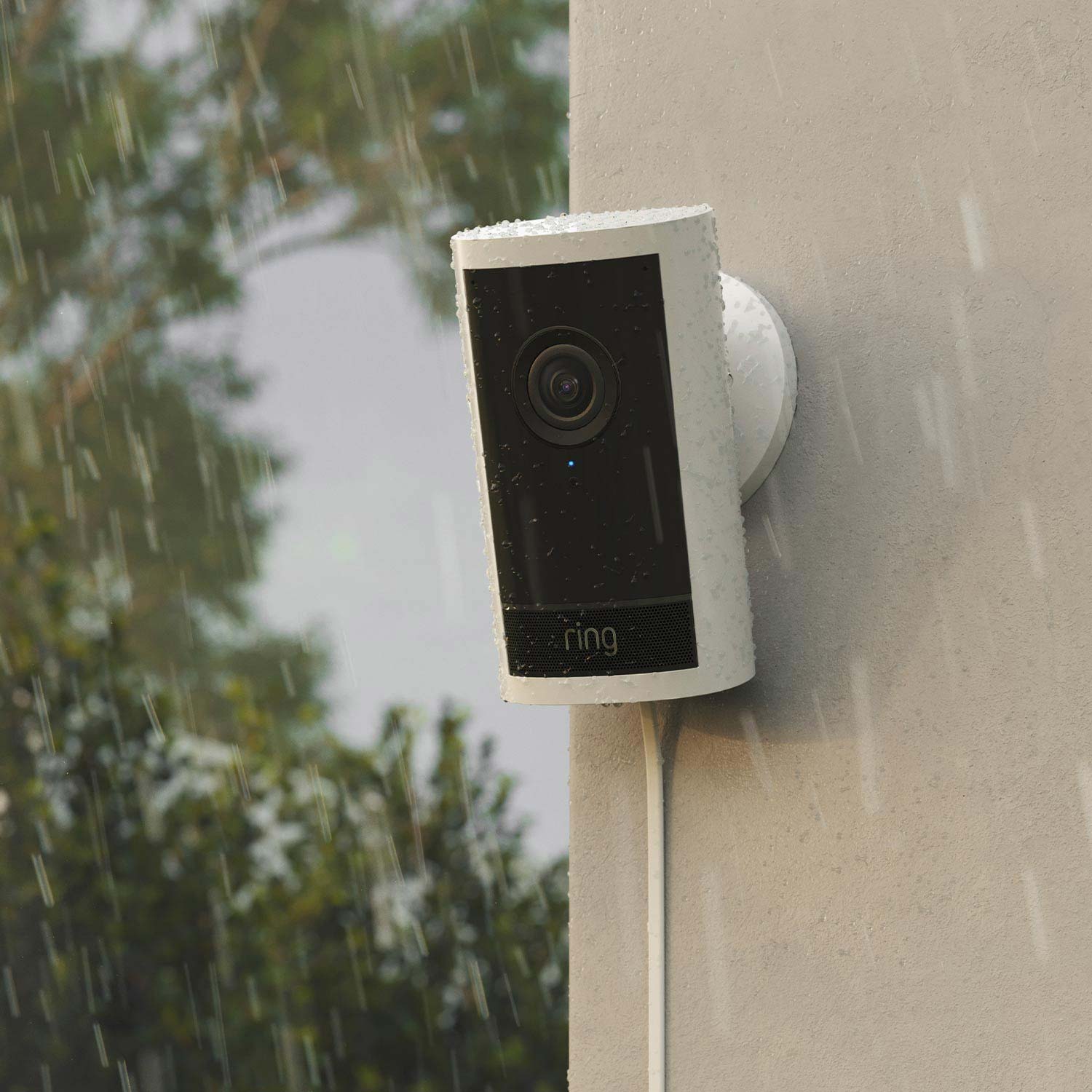 Outdoor Cam 4K Pro - White Ring Outdoor Cam 4K Pro mounted on a wall in the rain, water droplets visible, cable running down, trees blurred in background.