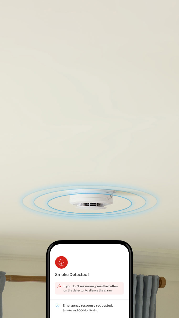 In a living room, a Kidde Smoke and CO Alarm is sounding on the cieling. A hand holds a mobile phone that shows a 