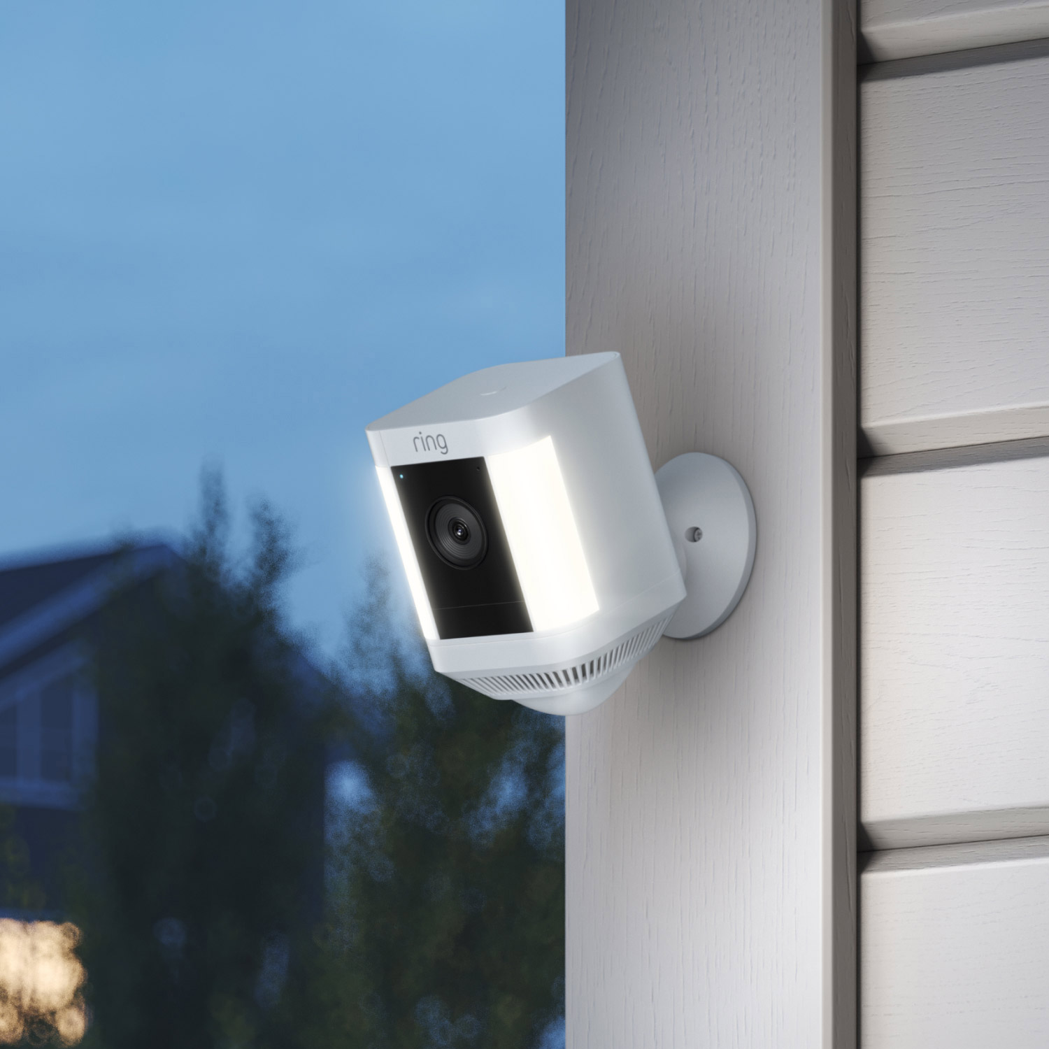 Battery Doorbell Pro + Spotlight Cam Plus Battery - Ring Spotlight Cam Plus mounted on an exterior wall, lit up at dusk with trees and houses in the background.