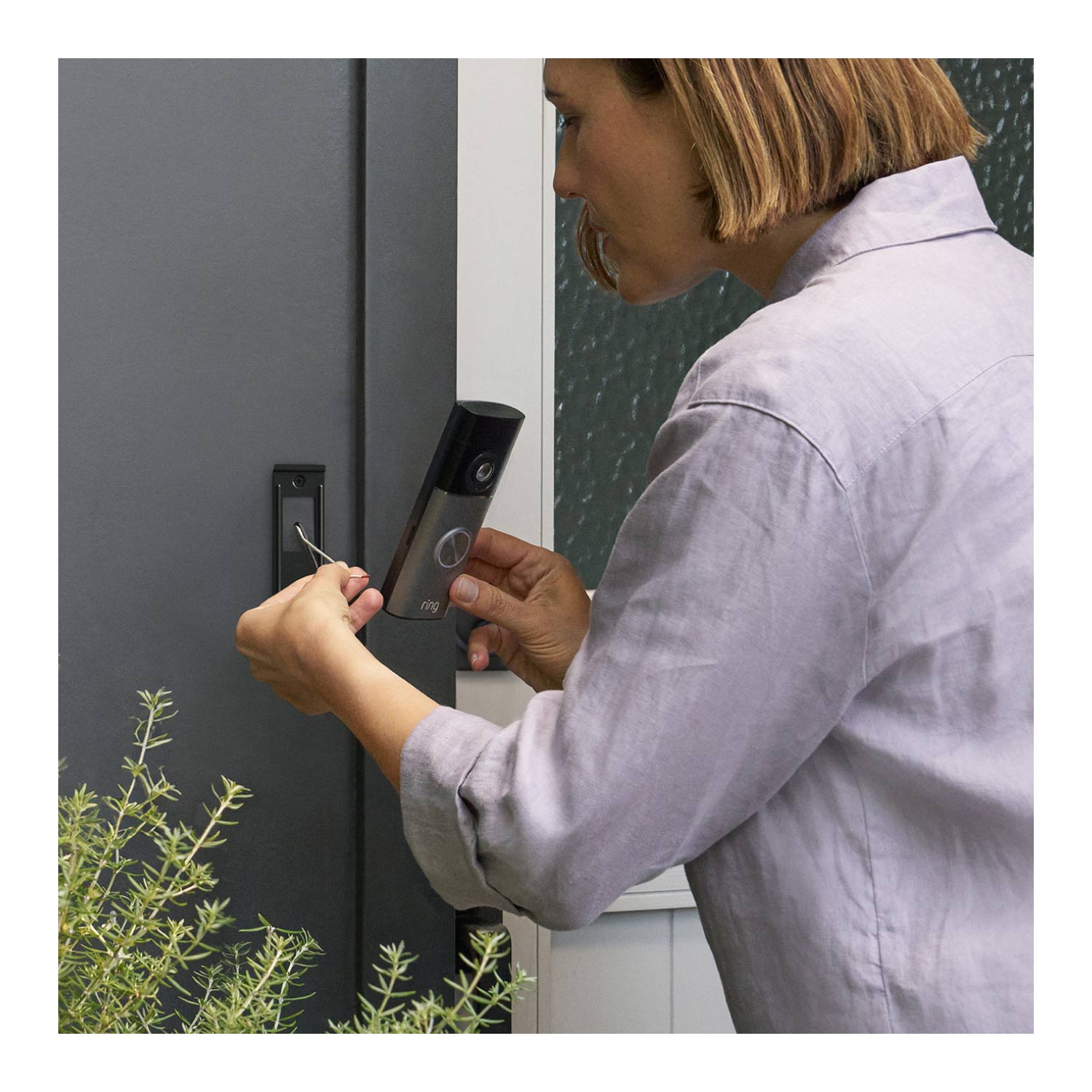 2-Pack Wired Doorbell Pro  (Newest Model) - Woman installing a Wired Doorbell Pro (2nd Gen), with their hands carefully mounting the device on a wall. Part of some greenery or plants is visible in the left bottom corner of the frame.